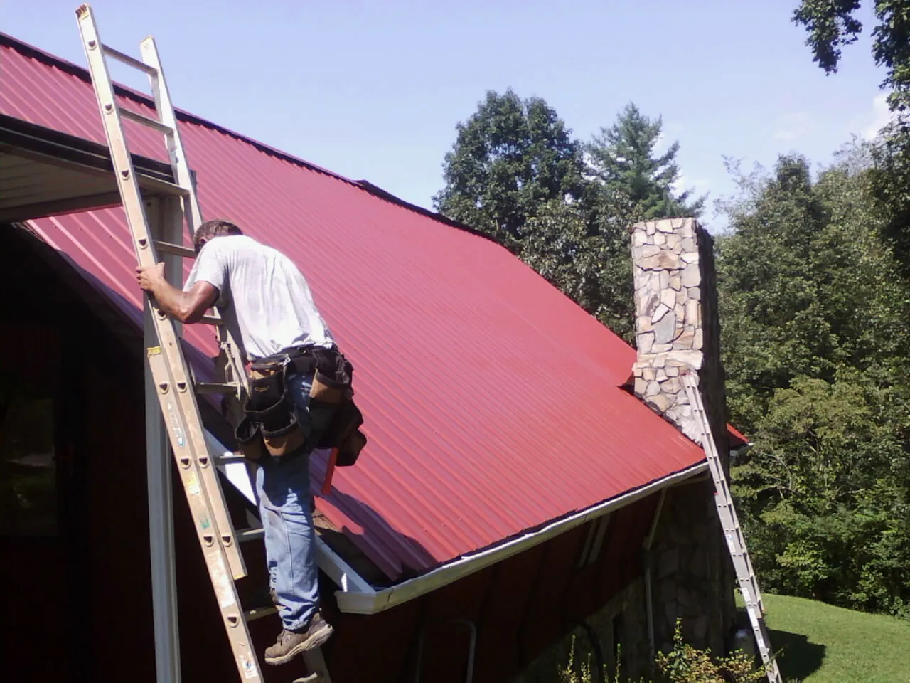 Roofer on a ladder beside a red metal roof and stone chimney — Soffit & Fascia Repair by Roofing Brothers in Corunna